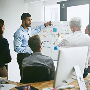 Middle Eastern man stands and points at plans on white board while multiracial colleagues discuss