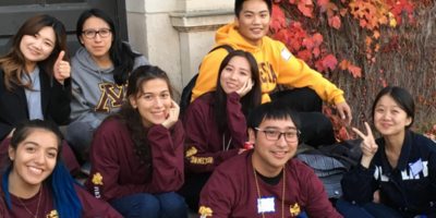 a group of IEP syudents wearing maroon and gold sit on the steps in front of a University building that is covered in maroon and gold leaves