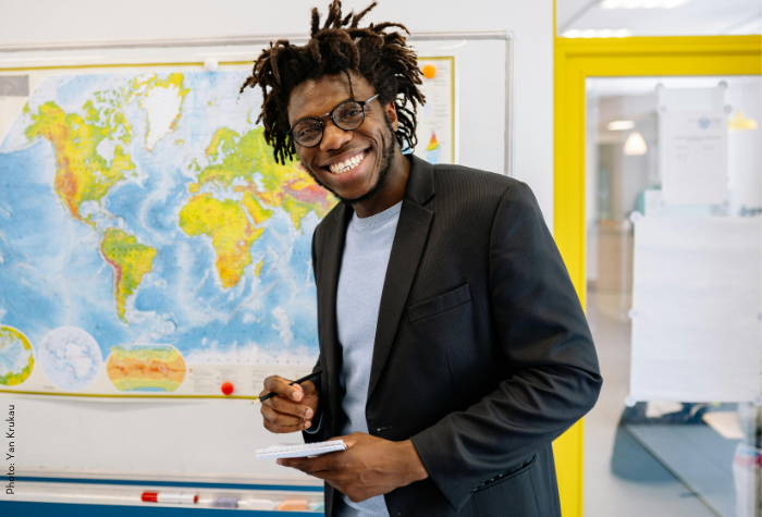 A high school instructor smiles for the camera while standing in his classroom in front of a large and colorful map; he hold a notepad and red pen