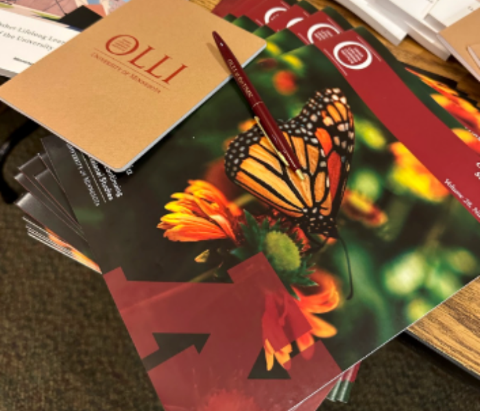OLLI publications and an OLLI pen with lush maroon and gold colors are strewn on a tabletop