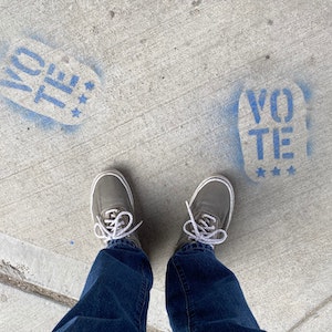 VOTE spray painted in blue on pavement with two shoes visible