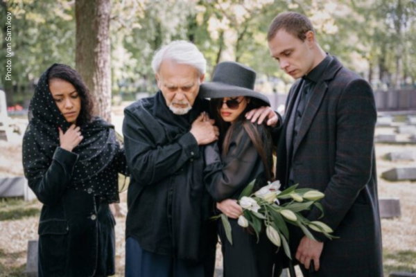 A family of four, all dressed in black mourning clothes, comfort one another while standing before the grave of a loved one.