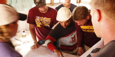A group of students wearing hard hats stand over blueprints while a supervisor gives instructions