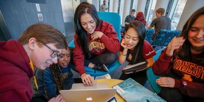Five students in Gopher shirts huddle around a laptop in a campus building lounge