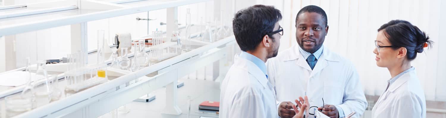 One white male, one Black male, and one Asian woman wearing white lab coats talk in a lab