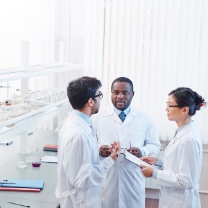 One white male, one Black male, and one Asian woman wearing white lab coats talk in a lab.