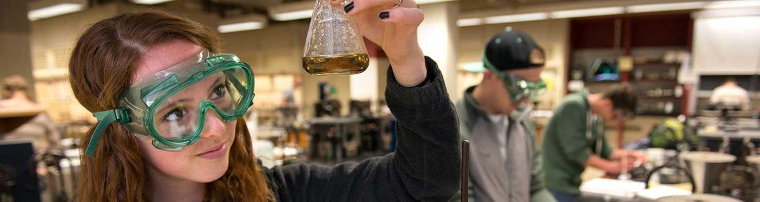 A high school student wears green lab googles and examines the contents of an Erlenmeyer flask, which she holds up to the light
