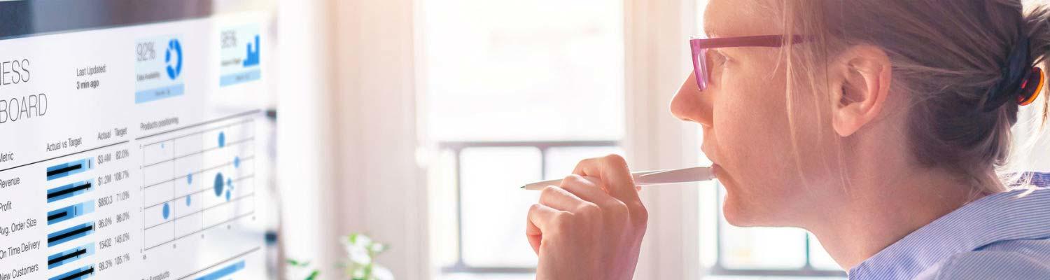 A person with light skin tone and blond hair pulled back into a ponytail rests a pen against their mouth while studying graphs on the computer monitor in front of them 