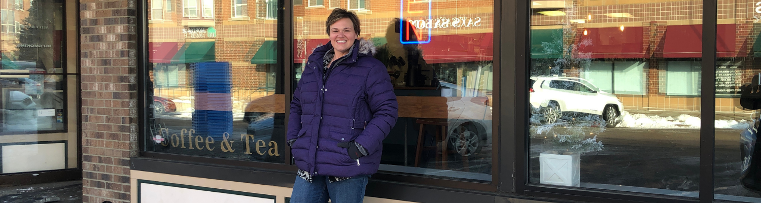 Tracy Shantz a white woman with short brown hair wearing a purple winter jacket stands in front of a cafe window reflecting the street