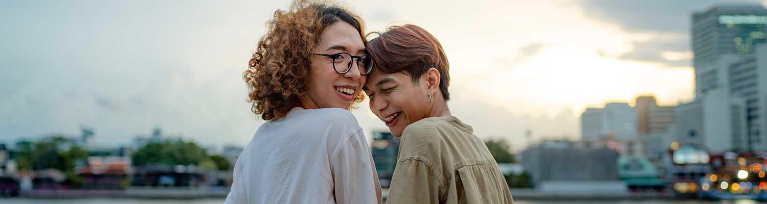 A young nonbinary couple embrace, looking over their shoulders at the camera, before an urban horizon.