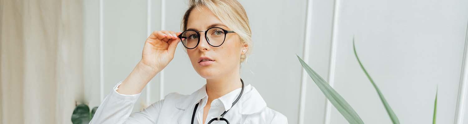 A woman doctor adjusts her glasses. She wears a stethoscope and holds a metal clipboard