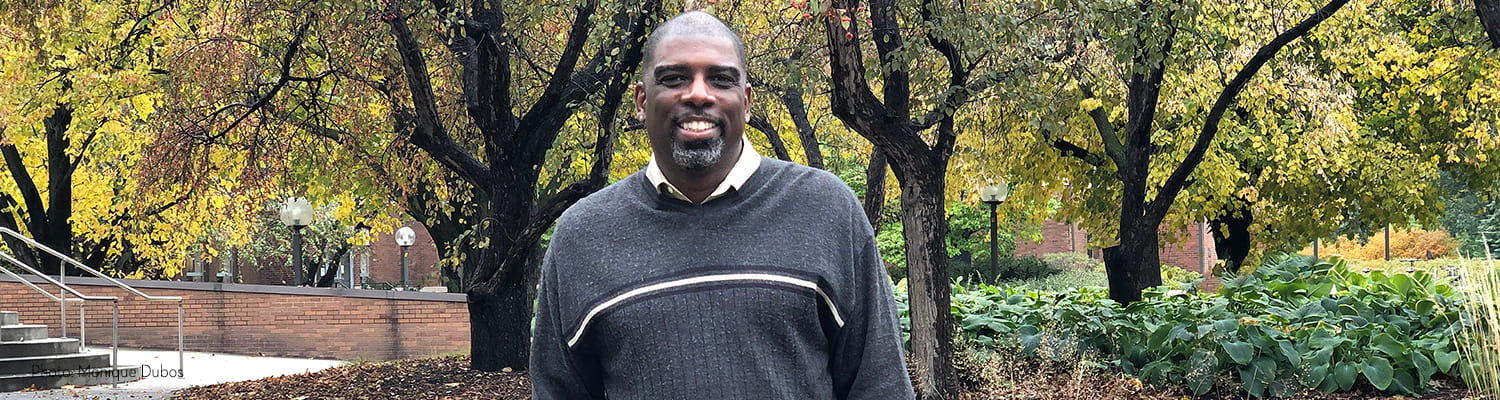 Black man wearing grey stiped sweater stands smiling with fall colored crab trees in background