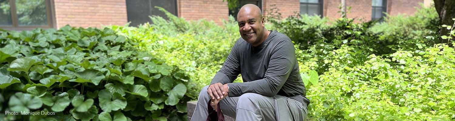 ITI Faculty Director Colin Miller, a Black man with a bald head, sitting on a stone bench surrounded by green shrubs, wearing grey shirt and pants, seen from the side, smiling at the camera