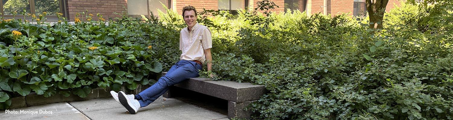 Luke Treiber sits on stone bench leaning back on his hands with legs outstretched and crossed in front of him and a garden of dense foliage and yellow flowers behind him