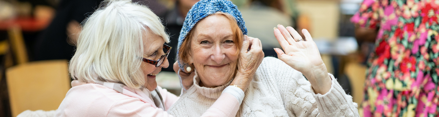 Two women in a knitting group play for the camera, with one woman putting a recently knit hat on the head of the other