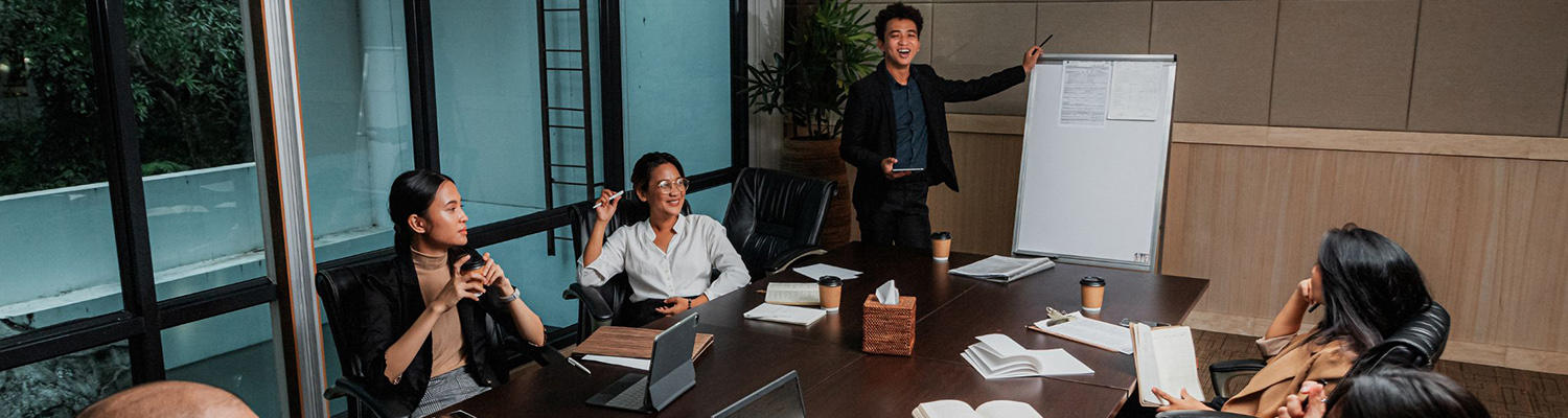 Young Asian man leads meeting at white board in front of table of other Asian professionals