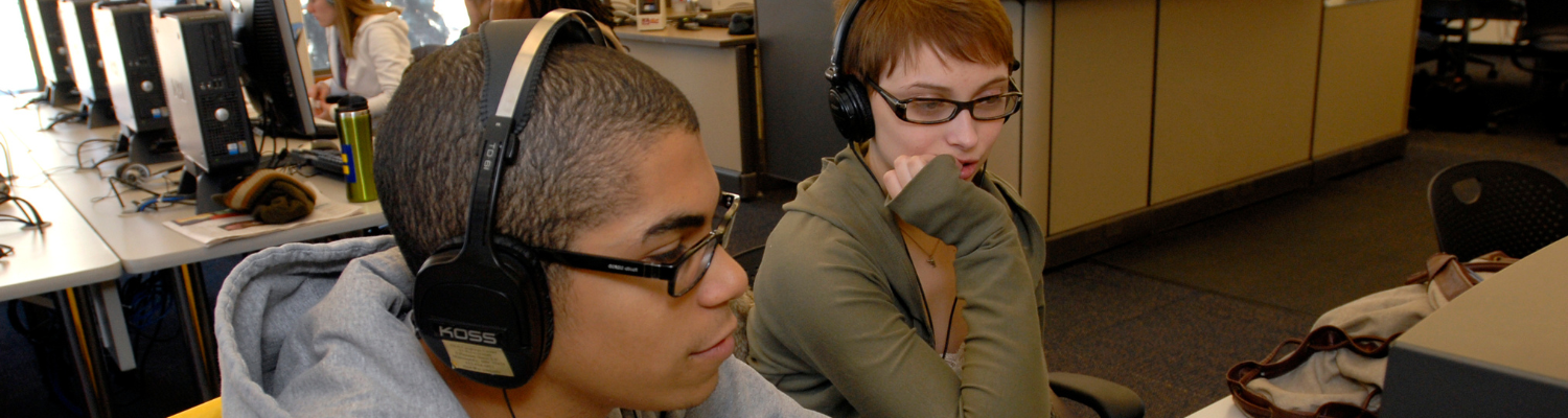 two young high school students wear headphones and sit at table intently watching a computer screen; other students may be seen working on computers in the background