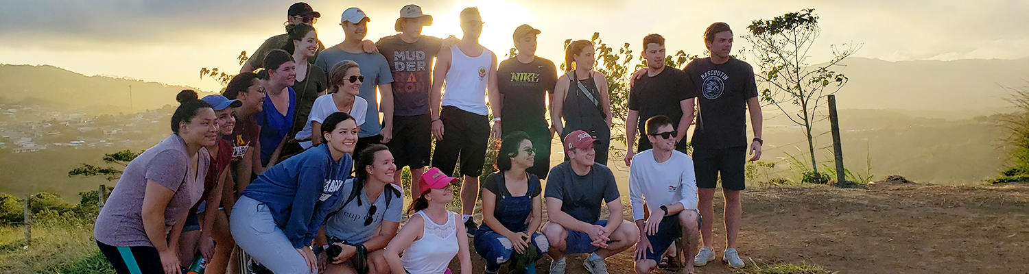 A group of students gather for a group photo on a hilltop in Costa Rica as the sun sets behind them