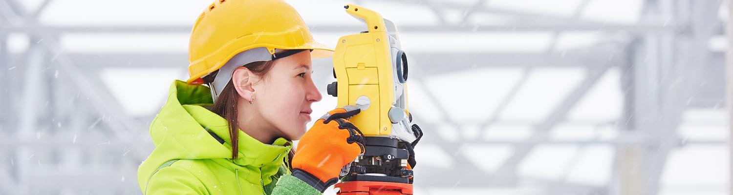 a white woman peers through a theodolite surveying a construction site