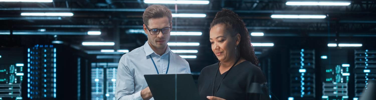 A white man and a Black woman read a laptop in the middle of a huge computer bank