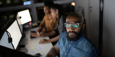 A Black man in thick rimmed glasses is lit by computer screen with two Black women looking at monitors in the background