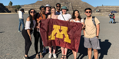 a diverse group of students hold a University of Minnesota flag in front of their group, standing in front of an archeological site in Mexico