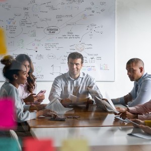 A group of professionals confer over a long table in a meeting room. Behind them, a whiteboard contains extensive notes.
